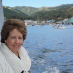 Women with brown hair and a white coat on the side of a boat with ocean and ships in the background.