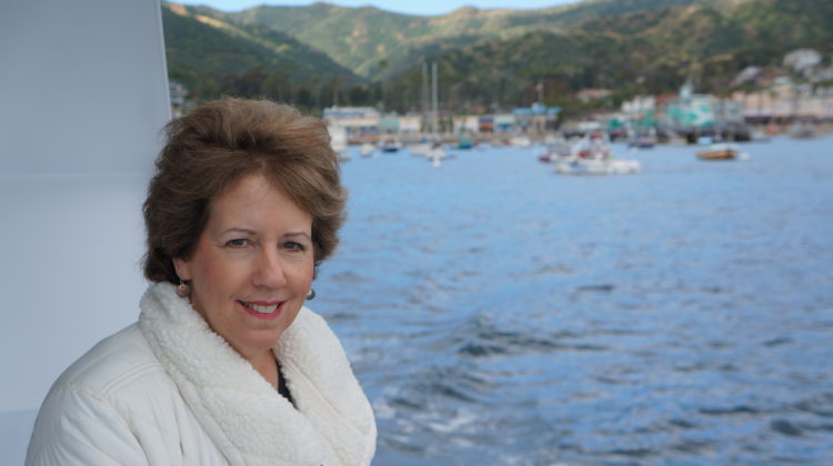 Women with brown hair and a white coat on the side of a boat with ocean and ships in the background.