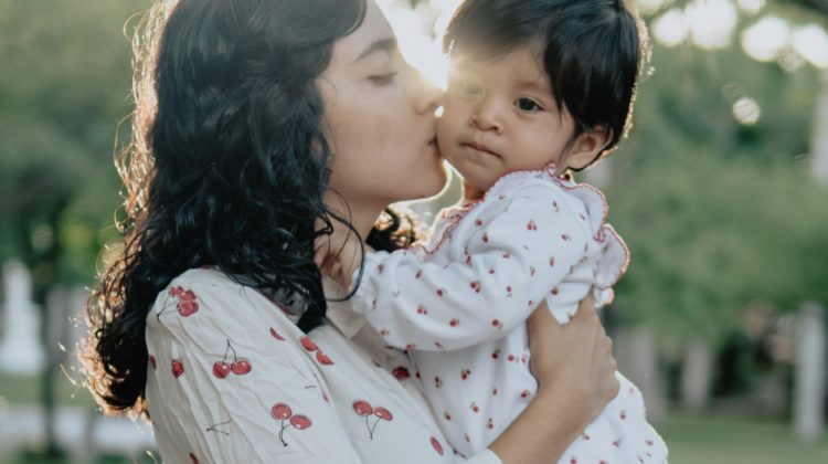 image of young woman with dark hair wearing a white blouse with cherries on it. She is holding and kissing a small baby