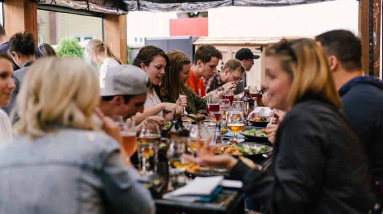 Large group of people sitting at a long table outside. There is many plates and glasses of beer and wine on the table.